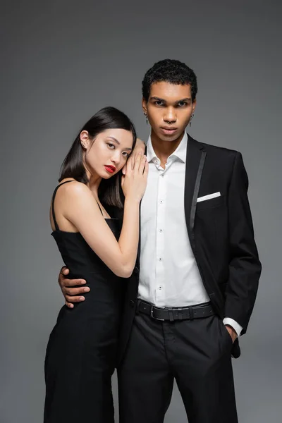 Elegant afrikanisch-amerikanischer Mann steht mit der Hand in der Tasche und umarmt sinnliche asiatische Frau mit roten Lippen isoliert auf grau — Stockfoto