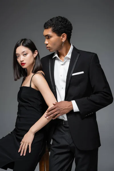 Africano americano homem no preto blazer olhando afastado perto asiático mulher no elegante vestido de cinta isolado no cinza — Fotografia de Stock
