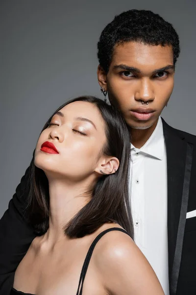 Elegante africano americano hombre mirando cámara cerca sensual asiático mujer con rojo labios y cerrado ojos aislado en gris - foto de stock