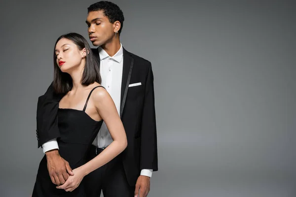 Stylische afrikanisch-amerikanische Mann im eleganten Anzug umarmt asiatische Frau posiert mit geschlossenen Augen isoliert auf grau — Stockfoto