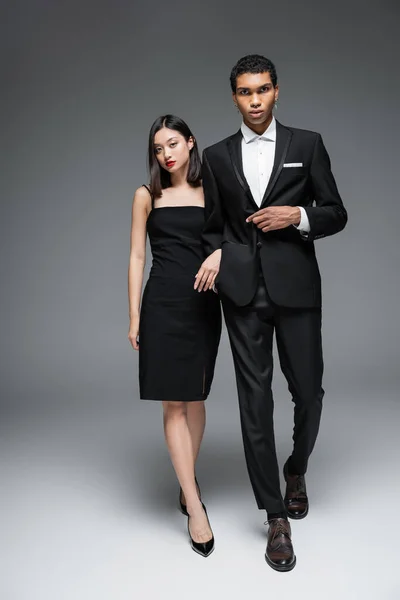 Full length of multiethnic couple in elegant formal wear walking and looking at camera on grey background — Stock Photo