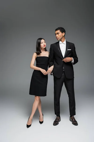 Полная длина межрасовой пары в черной элегантной одежде, смотрящей друг на друга на сером фоне — стоковое фото