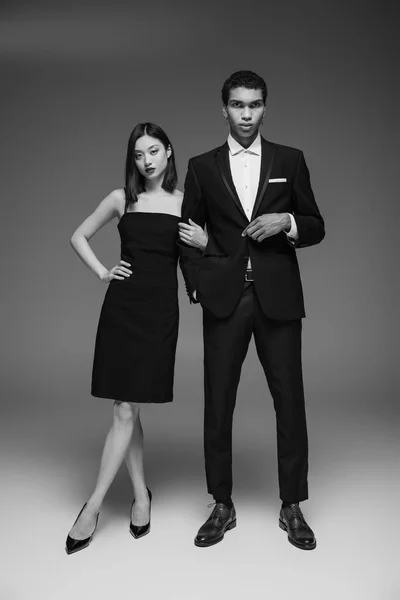 Comprimento total de homem americano africano em terno preto e mulher asiática em vestido elegante posando em fundo cinza — Fotografia de Stock