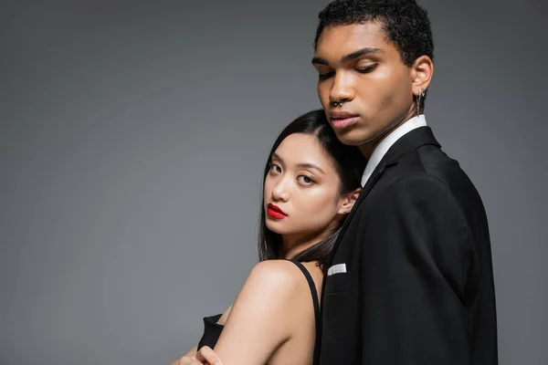 Encantadora mujer asiática con labios rojos apoyados en hombre afroamericano en chaqueta negra aislado en gris — Stock Photo