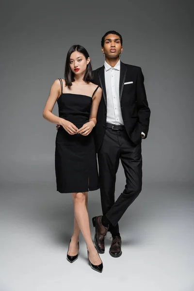 Full length of asian woman in black dress and african american man in stylish suit standing on grey background — Stock Photo