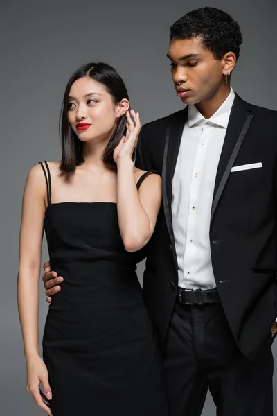 Afrikanisch-amerikanischer Mann im schwarzen Blazer umarmt Taille der eleganten asiatischen Frau lächelnd und wegschauend isoliert auf grau — Stockfoto
