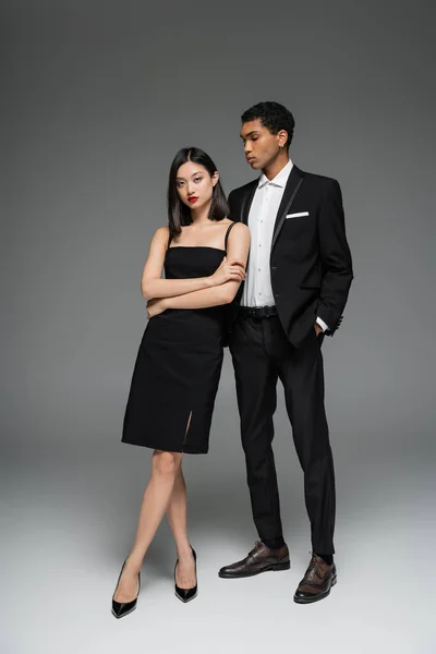 Longitud completa de la mujer asiática en vestido negro y afroamericano chico en elegante traje posando sobre fondo gris - foto de stock