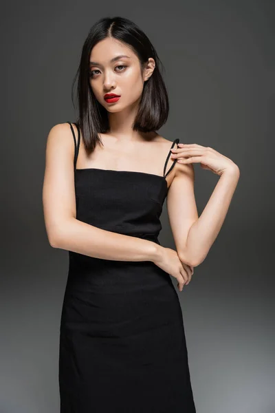 Encantadora mujer asiática en negro elegante vestido mirando a la cámara sobre fondo gris - foto de stock