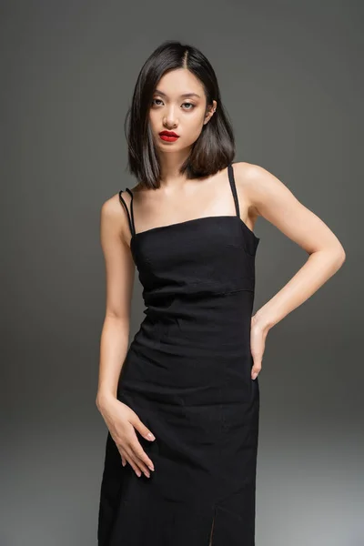 Morena asiática mulher no preto elegante vestido posando com a mão no quadril no fundo cinza — Fotografia de Stock