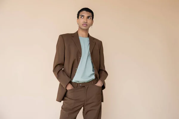 Hombre afroamericano de moda en traje marrón cogido de la mano en bolsillos y mirando hacia otro lado aislado en beige - foto de stock