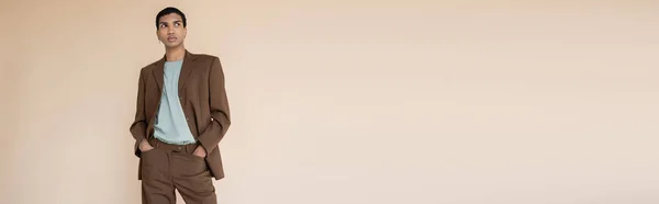 Hombre afroamericano de moda en traje marrón mirando hacia otro lado mientras está de pie con las manos en bolsillos aislados en beige, bandera - foto de stock
