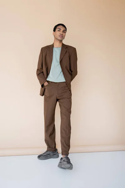 Intera lunghezza dell'uomo afroamericano in abito marrone in piedi con mano in tasca su sfondo beige — Foto stock