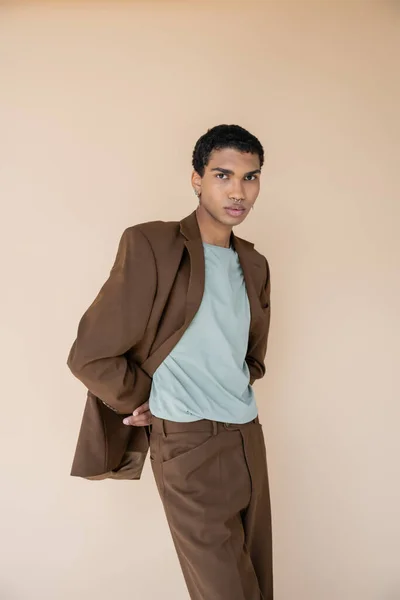 Élégant homme afro-américain en blazer marron regardant la caméra tout en posant avec les mains derrière le dos isolé sur beige — Photo de stock