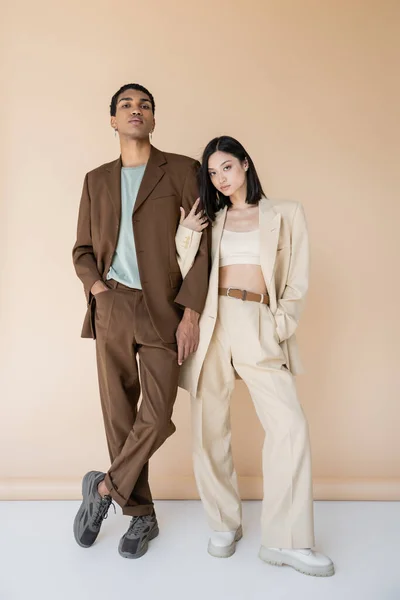 Full length of multiethnic couple in trendy pantsuits posing with hands in pockets on beige background — Stock Photo