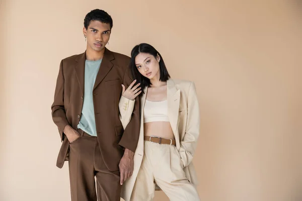 Jovem casal multiétnico na moda pantsuits posando com as mãos em bolsos isolados no bege — Fotografia de Stock