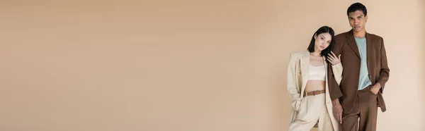 Jeune couple interracial en costumes tendance debout avec les mains dans les poches isolées sur beige, bannière — Photo de stock