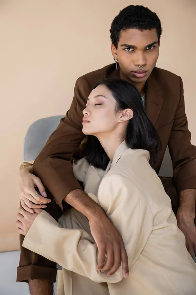 Young african american man looking at camera near fashionable asian woman with closed eyes on beige background — Stock Photo