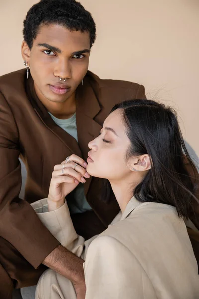 Stylish african american guy with piercing looking at camera near young asian woman with closed eyes isolated on beige — Stock Photo