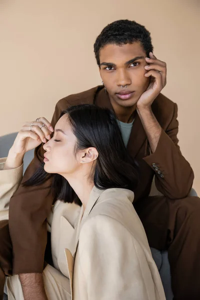 Stylish african american man in brown suit looking at camera near sensual asian woman with closed eyes isolated on beige — Stock Photo