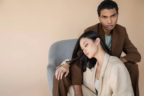 Hombre afroamericano con estilo mirando a la cámara cerca sensual mujer asiática en traje de marfil aislado en beige - foto de stock
