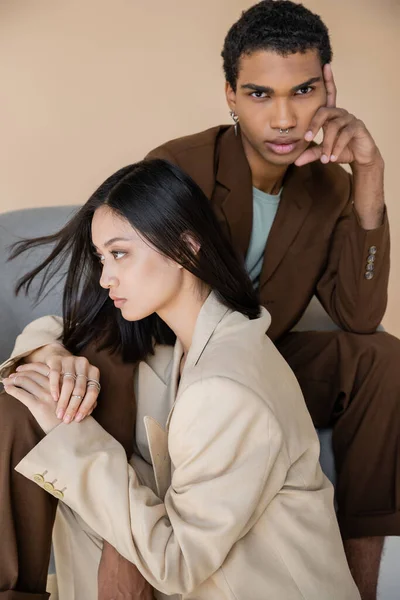Hombre afroamericano elegante sentado en sillón y mirando a la cámara cerca de la mujer asiática en chaqueta de marfil sobre fondo beige - foto de stock