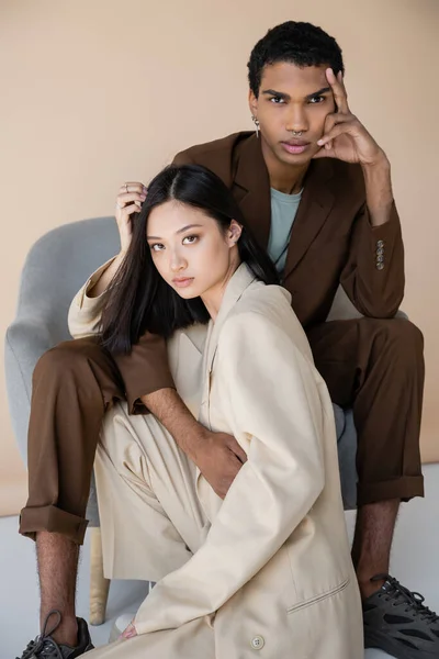 Fashionable interracial models in trendy pantsuits looking at camera near armchair on beige background — Stock Photo