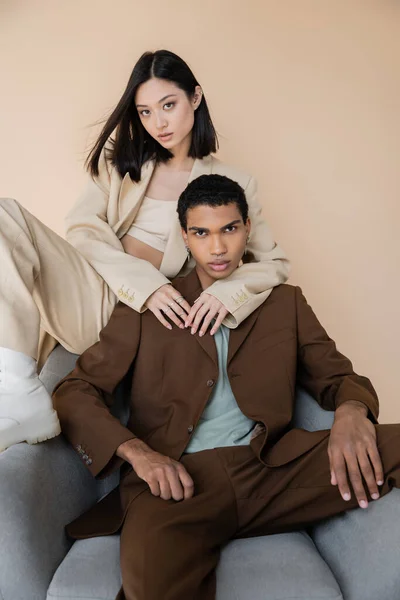 Sensual asian woman embracing african american man in trendy suit sitting in armchair isolated on beige — Stock Photo