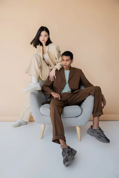 Comprimento total de jovem casal interracial na moda pantsuits posando em poltrona no fundo bege — Fotografia de Stock