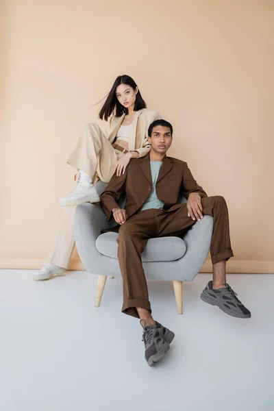 Longitud completa de pareja interracial en elegantes trajes de pantalón posando cerca de sillón sobre fondo beige - foto de stock