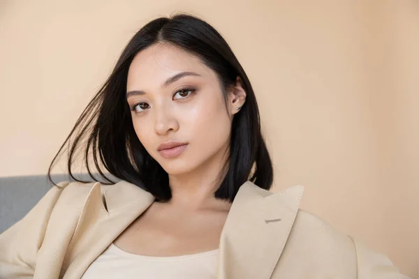 Portrait of brunette asian woman in ivory blazer looking at camera on beige background — Stock Photo