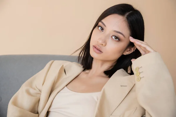 Young asian woman in blazer holding hand near face and looking at camera on beige background — Stock Photo