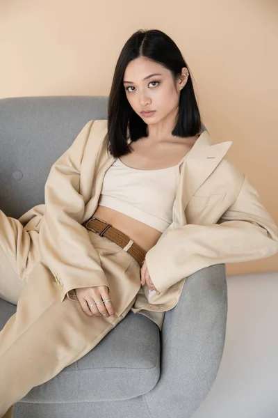 Sensual asian woman in stylish pantsuit sitting in armchair and looking at camera on beige background — Stock Photo