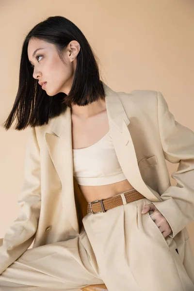 Bruna donna asiatica in elegante pantalone posa con mano in tasca e guardando lontano su sfondo beige — Foto stock