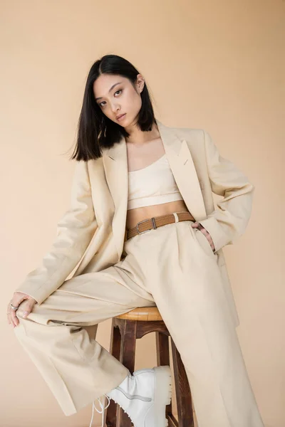 Stylish asian woman in pantsuit holding hand in pocket while posing on stool isolated on beige — Stock Photo