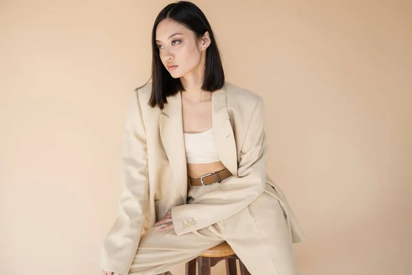 Trendy asian woman in ivory suit sitting on stool and looking away isolated on beige — Stock Photo