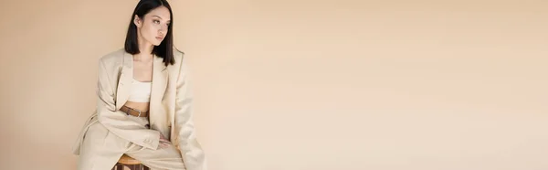 Brunetta donna asiatica in elegante abito avorio seduto su sgabello e guardando lontano isolato sul beige, banner — Foto stock