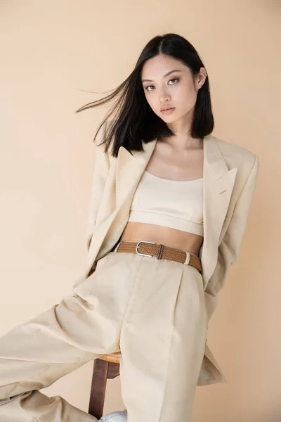 Fashionable asian woman in pantsuit posing near stool and looking at camera isolated on beige — Stock Photo