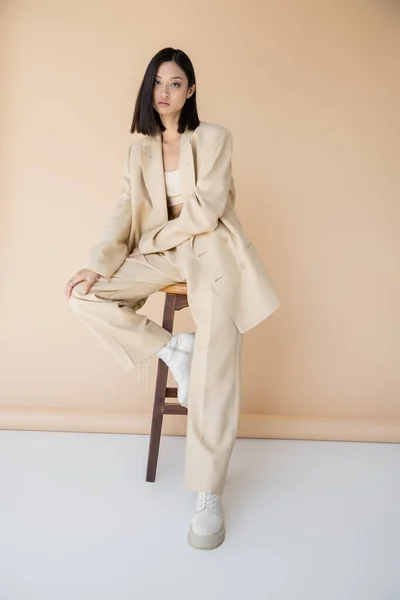 Brunette asian woman in stylish pantsuit and boots sitting on stool on beige background — Stock Photo