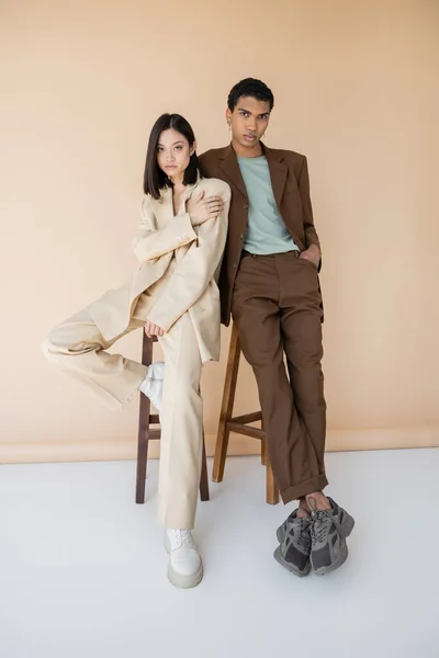Longitud completa de la joven pareja multiétnica en trajes elegantes posando cerca de taburetes sobre fondo beige - foto de stock