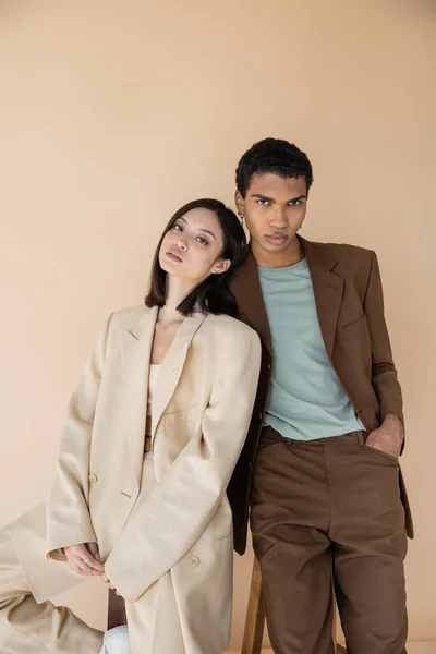 Interracial couple in fashionable blazers looking at camera isolated on beige — Stock Photo
