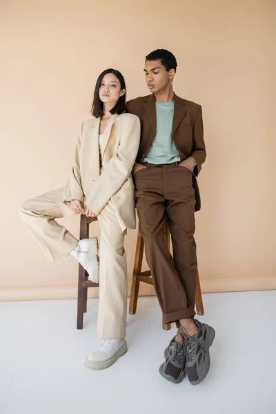 Longitud completa de pareja interracial en ropa de moda posando cerca de taburetes sobre fondo beige - foto de stock
