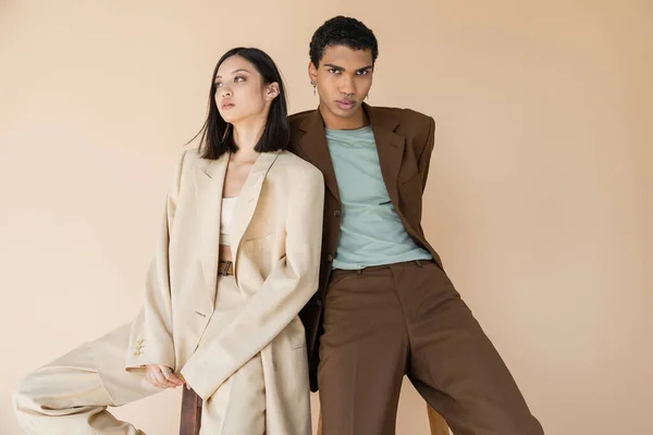 Alla moda afroamericano uomo in abito marrone guardando fotocamera vicino bruna modello asiatico isolato su beige — Foto stock