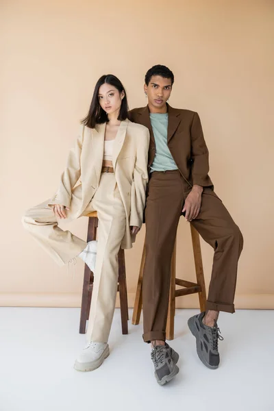 Longitud completa de la pareja multiétnica en elegantes trajes de pantalón posando cerca de taburetes sobre fondo beige - foto de stock