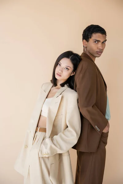 Casal interracial na moda em ternos de pé de volta para trás com as mãos em bolsos isolados no bege — Fotografia de Stock