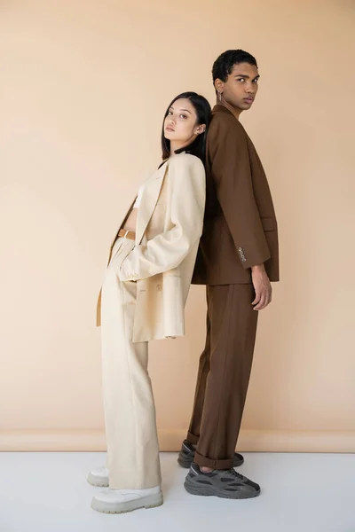 Full length of asian woman and african american man in trendy pantsuits standing back to back on beige background — Stock Photo