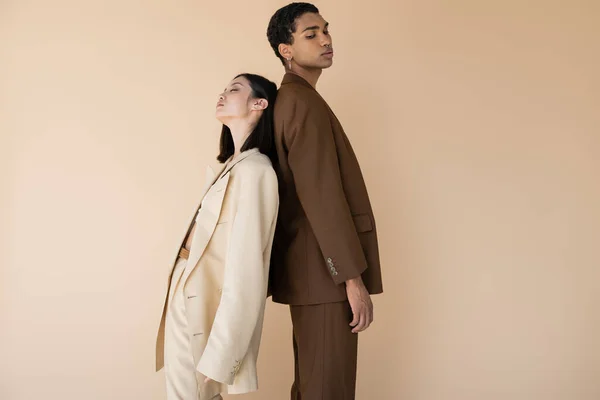 Sensual mujer asiática con los ojos cerrados de pie espalda con espalda con hombre afroamericano en traje marrón aislado en beige - foto de stock