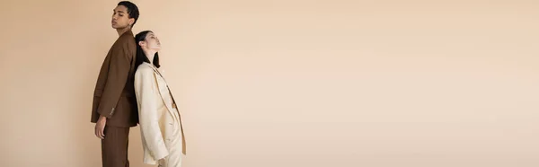 Joven pareja interracial en traje de moda posando espalda con espalda aislado en beige, pancarta — Stock Photo