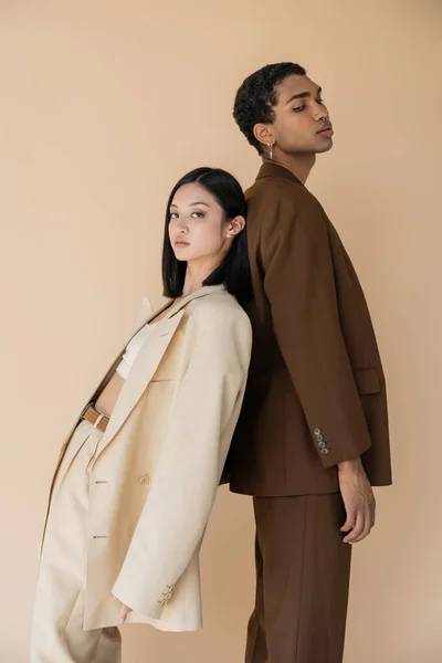Joven mujer asiática en pantsuit de pie espalda con espalda con el hombre afroamericano de moda y mirando a la cámara aislado en beige - foto de stock