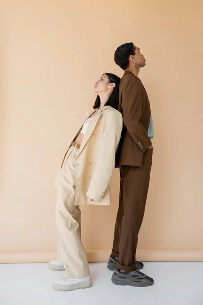 Full length of interracial couple in stylish pantsuits posing back to back on beige background — Stock Photo