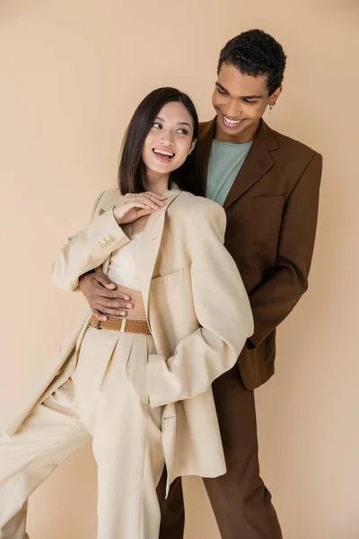 Stylish african american man embracing carefree asian woman in ivory suit isolated on beige — Stock Photo